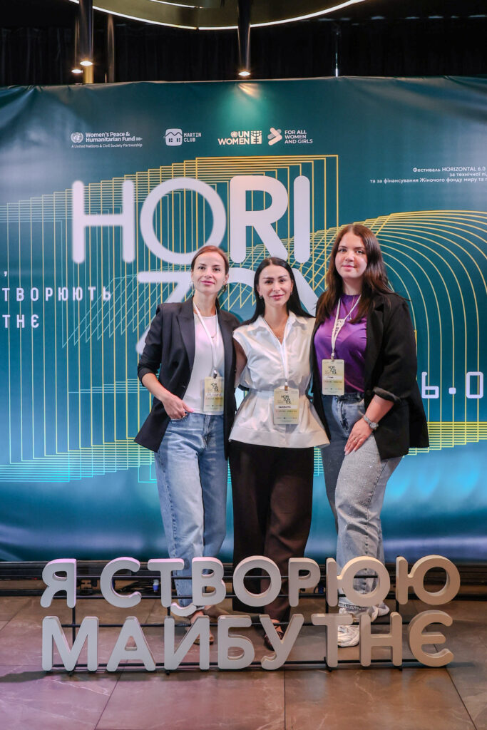 Представниці ГО «МАРТІН-клуб» та партнерської організації на конференції «Жінки, які створюють майбутнє» на фоні банера з логотипами UN Women та ГО «МАРТІН-клуб».